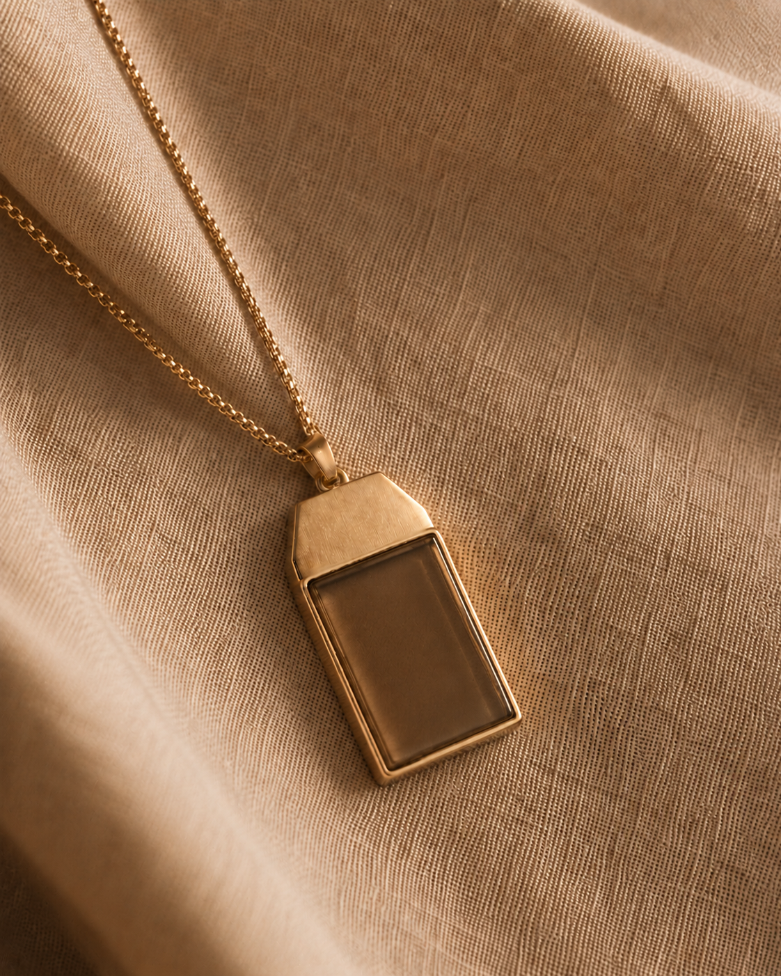 The Somatag pendant — a brushed-gold tag with a tiger's-eye stone face, resting on warm folded linen